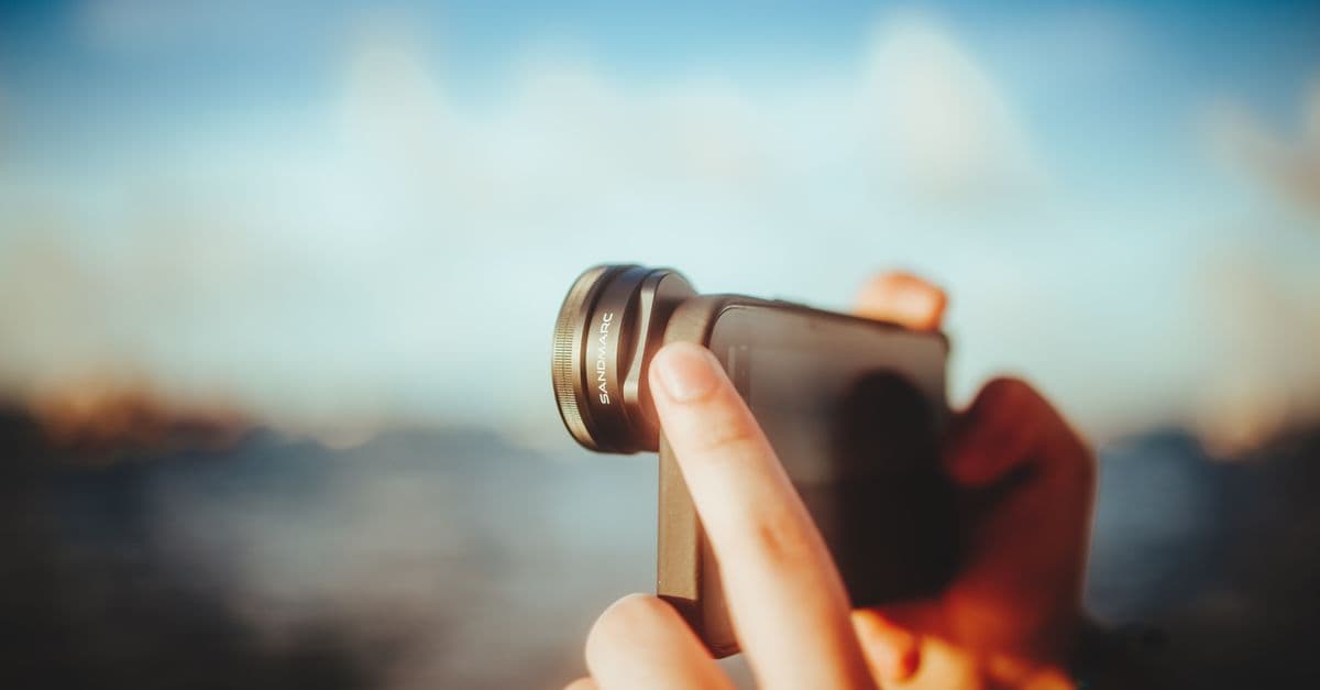 A close-up of hands using a smartphone with a lens to capture outdoor images, against a blurred background.